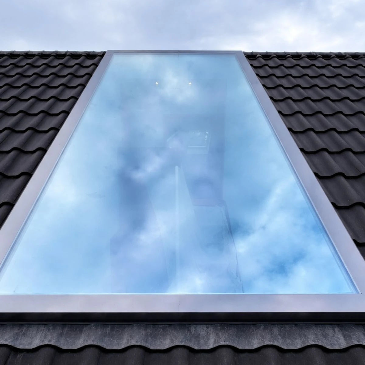Schrägdachfenster von außen: Glasfläche spiegelt den bewölkten Himmel, schmaler heller Rand, dunkle Dachziegel ringsum